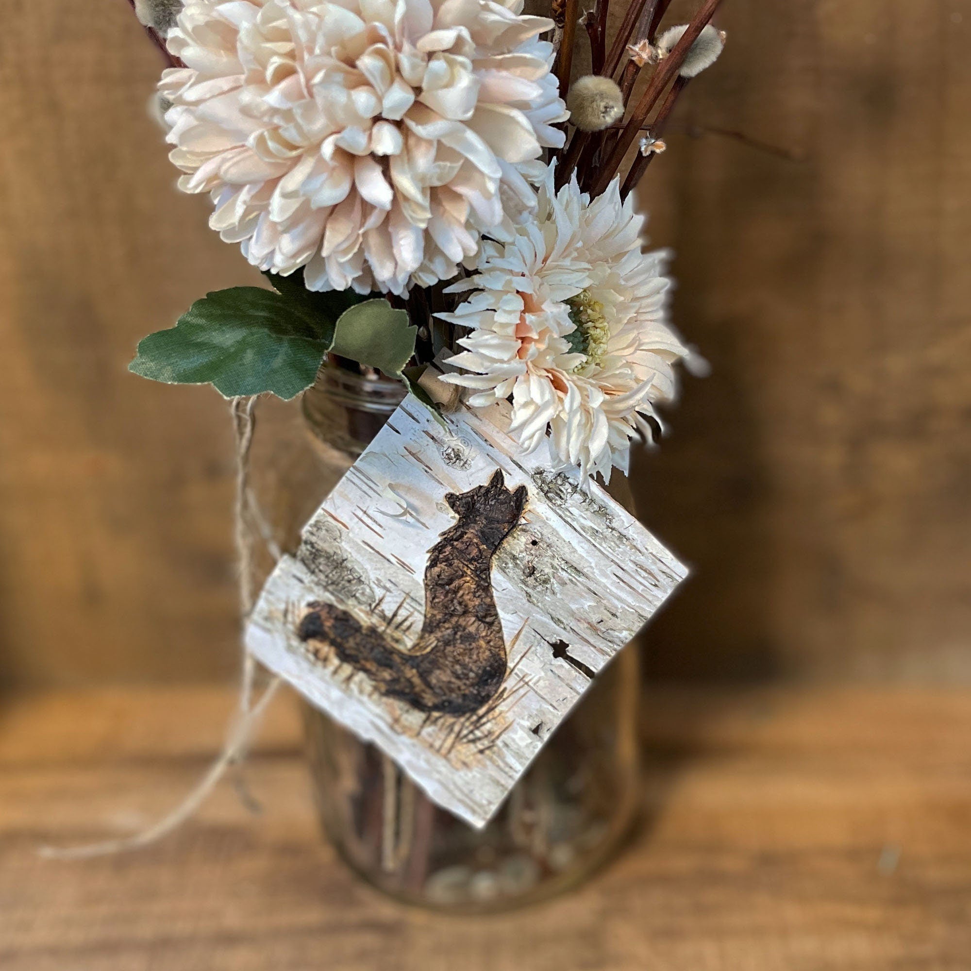 Decorative vase with flowers and a birch tag featuring a fox on a wooden surface