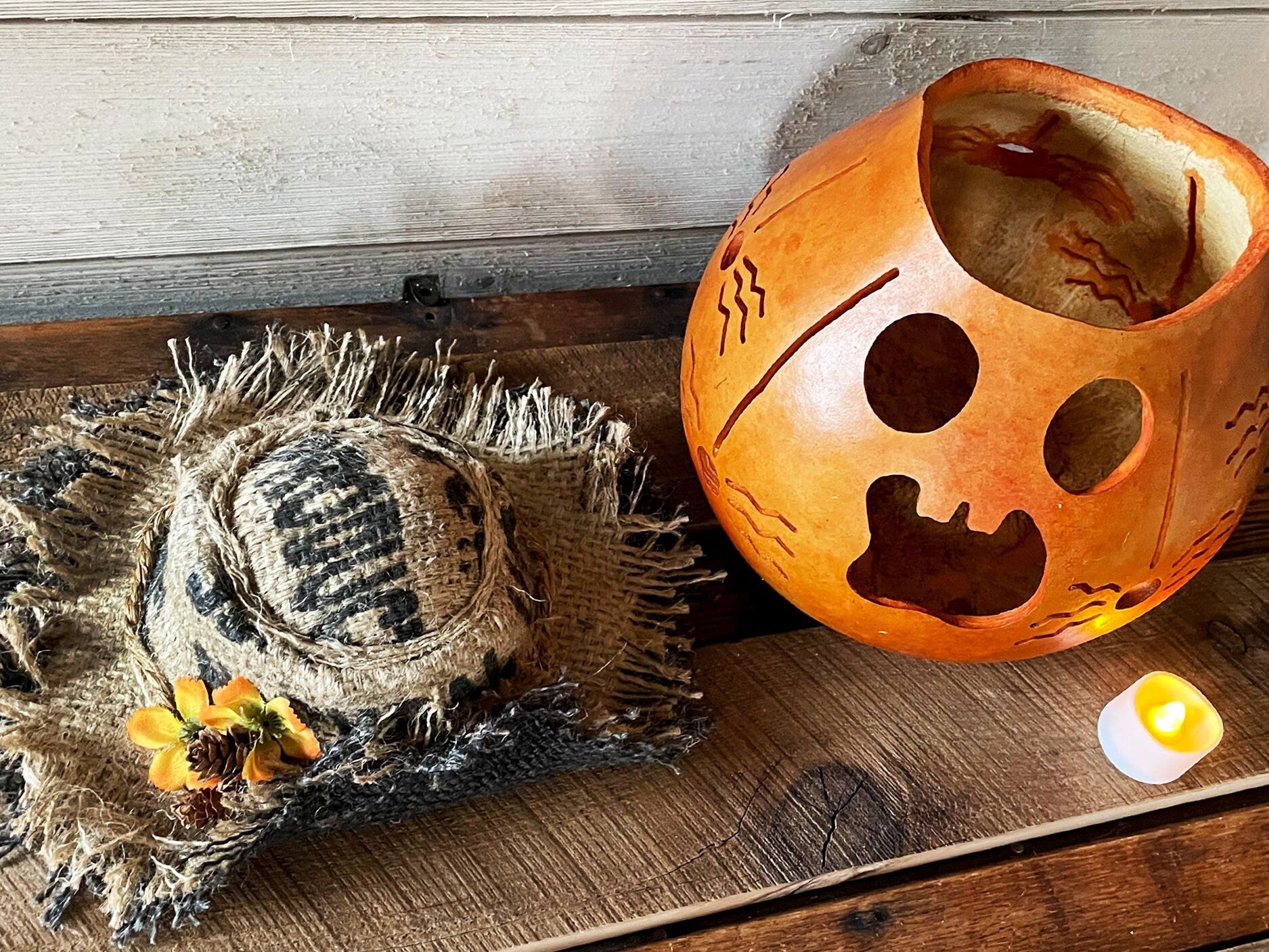 Decorative pumpkin with carved face on a wooden surface, next to a burlap hat and small candle.