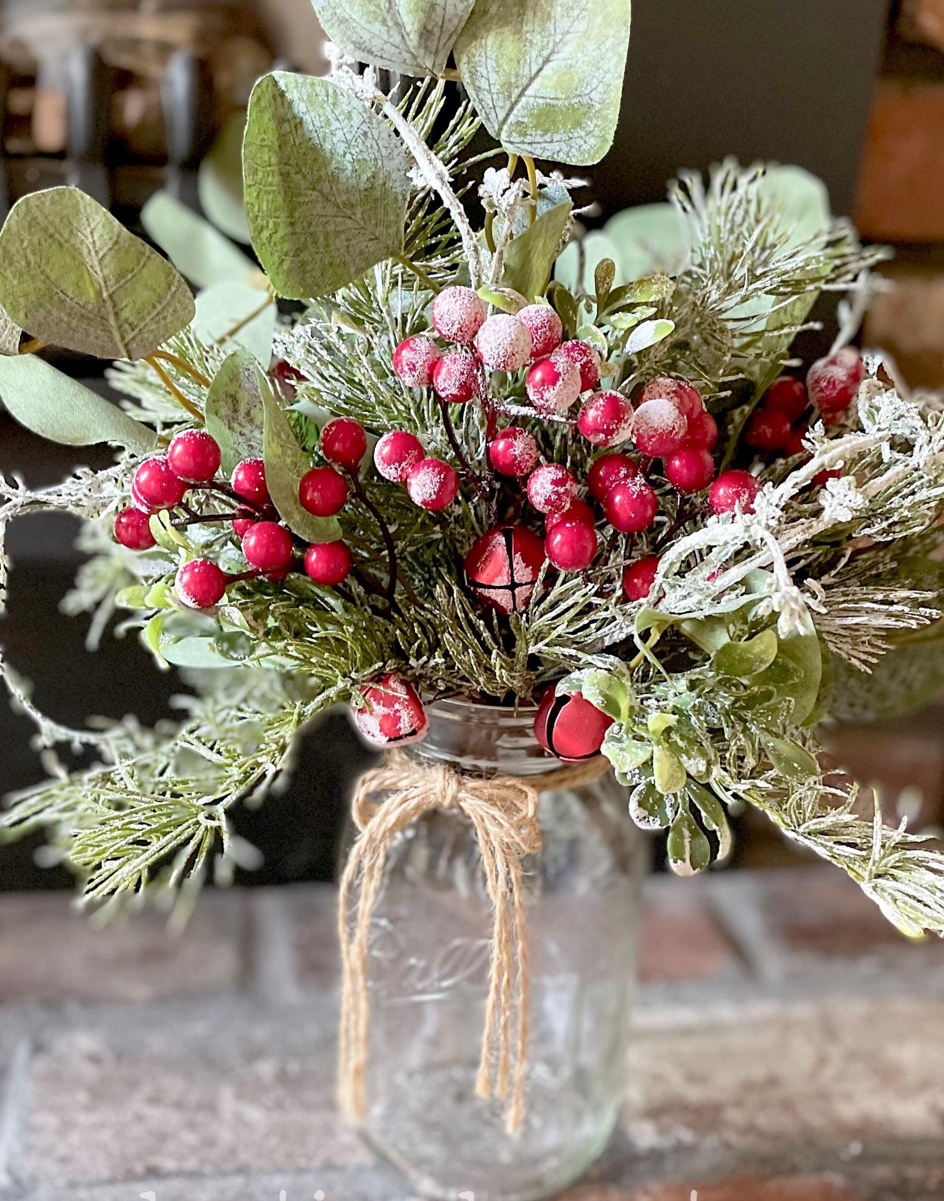 Decorative arrangement of greenery and red berries in a clear jar on a wooden surface.