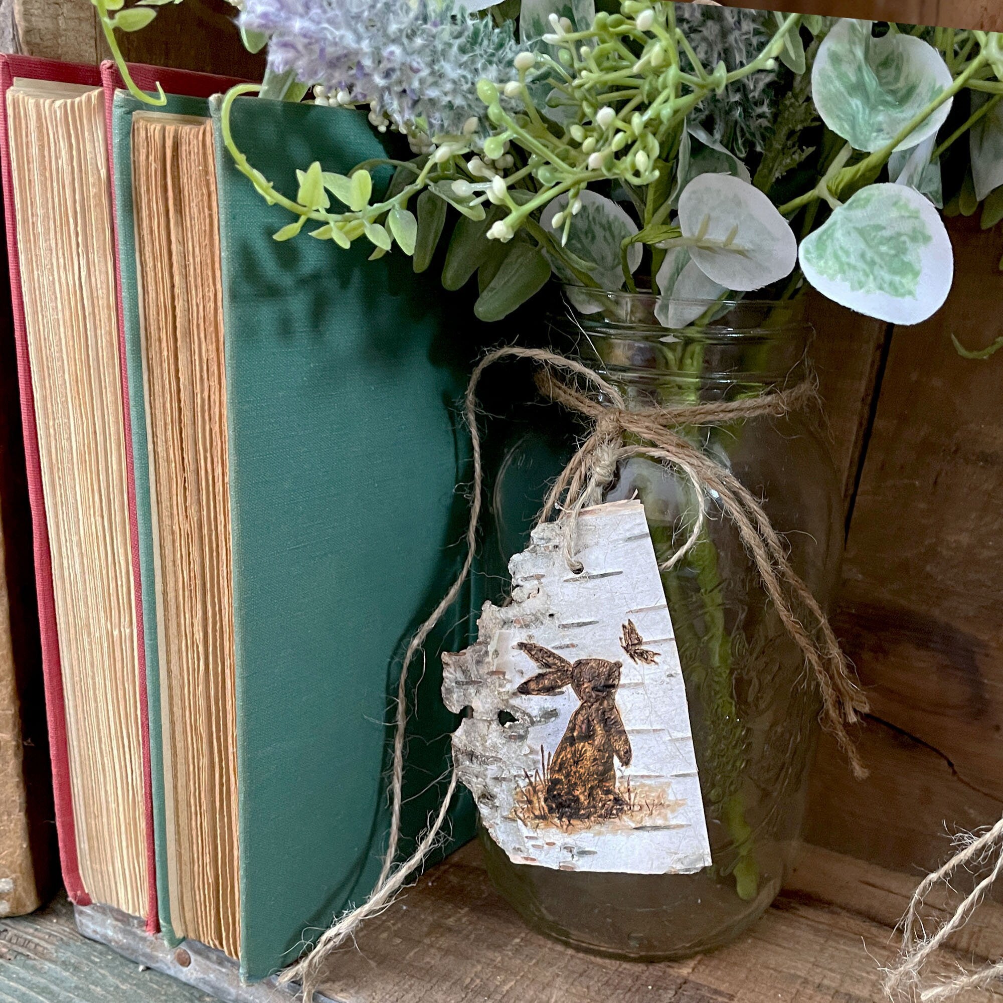 Mason jar with a rabbit tag, greenery, and books on a wooden surface