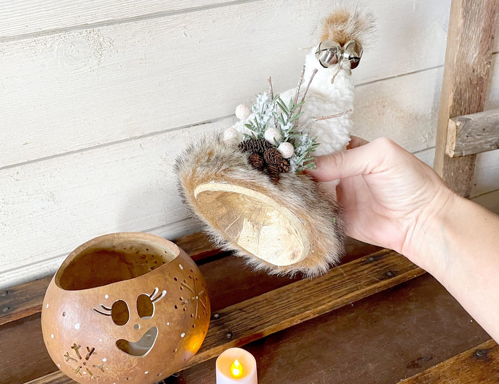 Decorative item with fur and floral elements held by a hand, next to a decorative pot on a wooden surface.