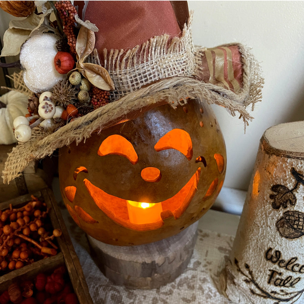 Decorative pumpkin with a face carved into it, wearing a burlap hat, on a table with fall-themed decorations.