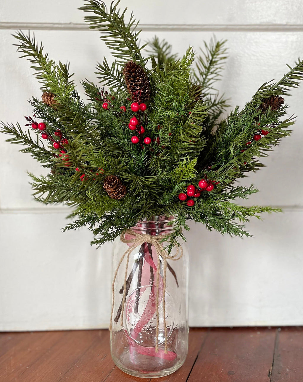 Bouquet of realistic pin and Fraser fur greenery with red berries in a clear glass jar on a wooden surface.