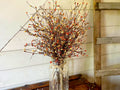 Decorative arrangement of branches with small orange berries in a clear glass jar on a wooden surface.