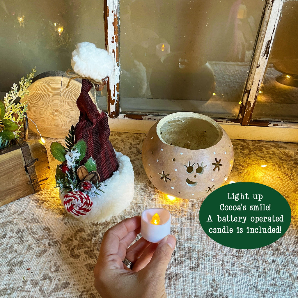 Decorative setup with a small Santa Claus figure, a ceramic pot with a face design, and a hand holding a battery-operated candle.
