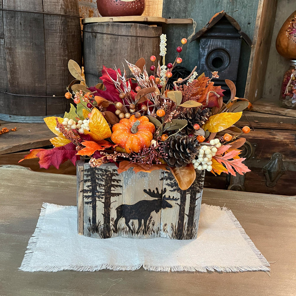 Decorative fall arrangement with pumpkins and leaves in a container with moose design on a wooden surface.
