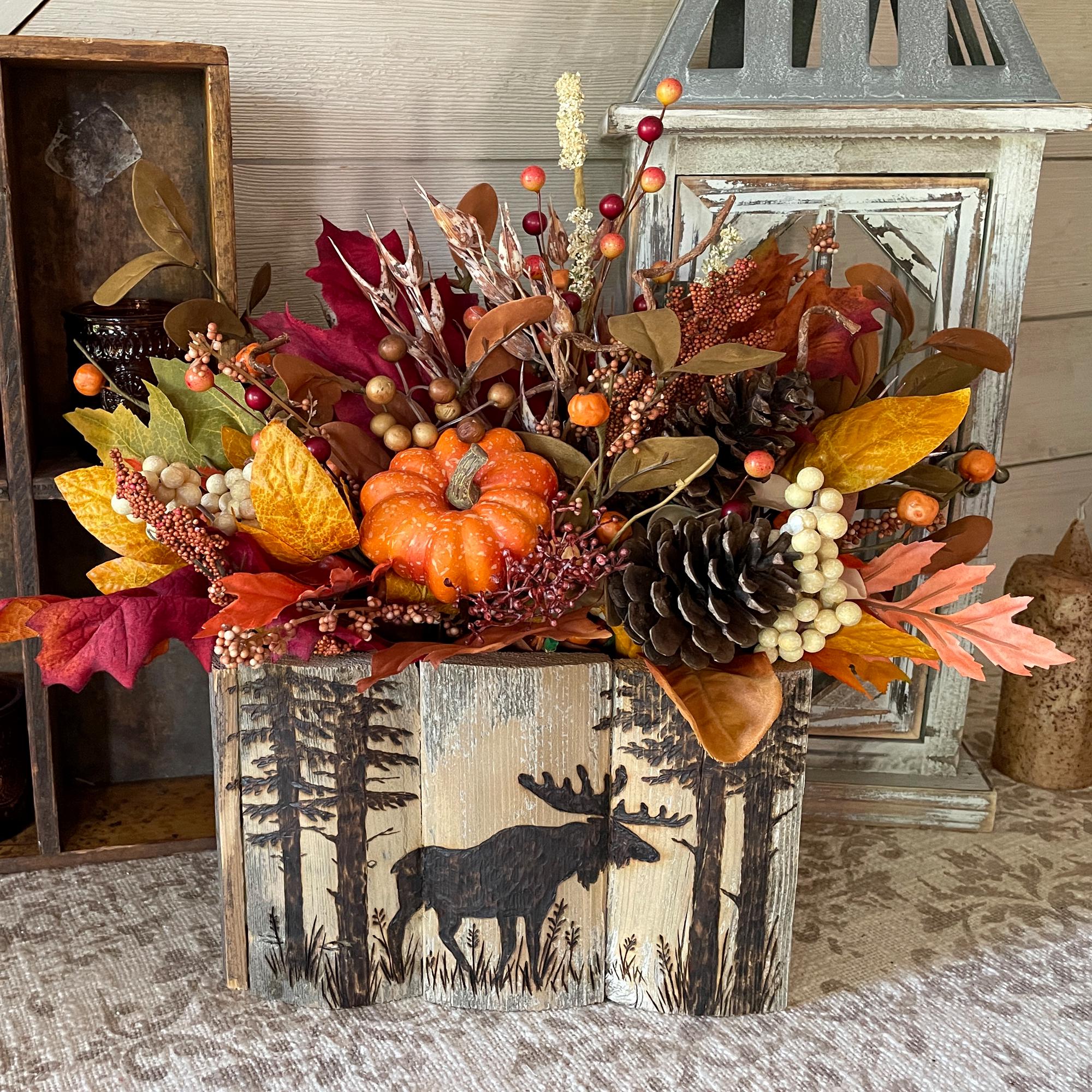 Decorative fall arrangement with pumpkins, leaves, and pinecones on a wooden block with a moose design.