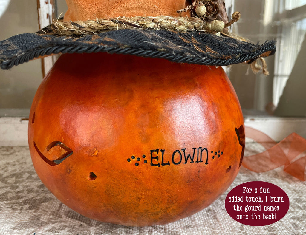 Decorative gourd with a face and 'ELOWIN' name, placed on a textured surface.