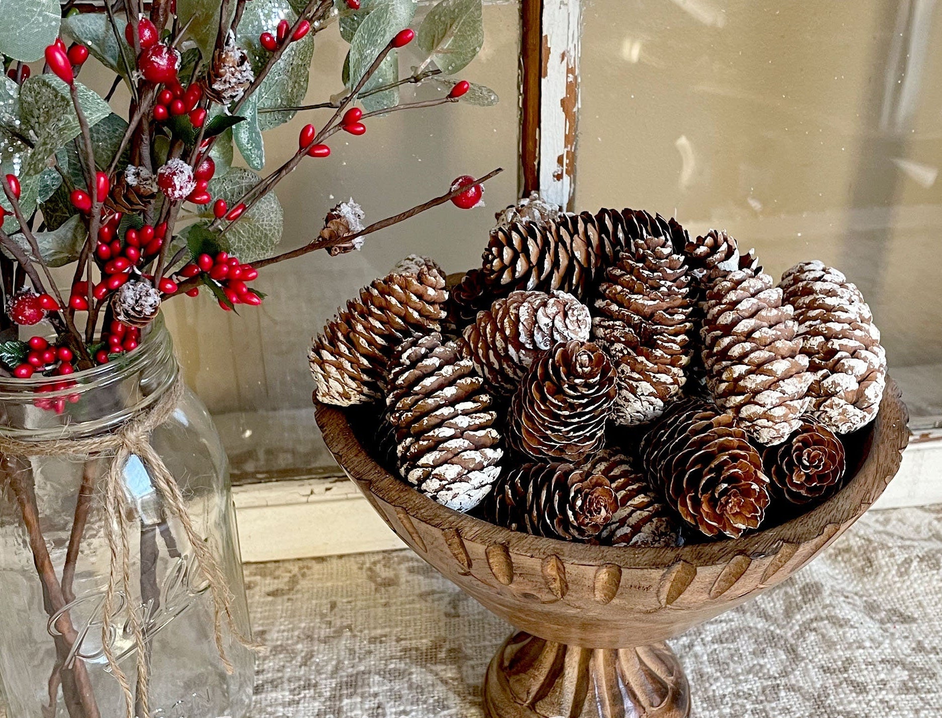 Decorative bowl with pinecones next to a jar with red berries on a textured surface.
