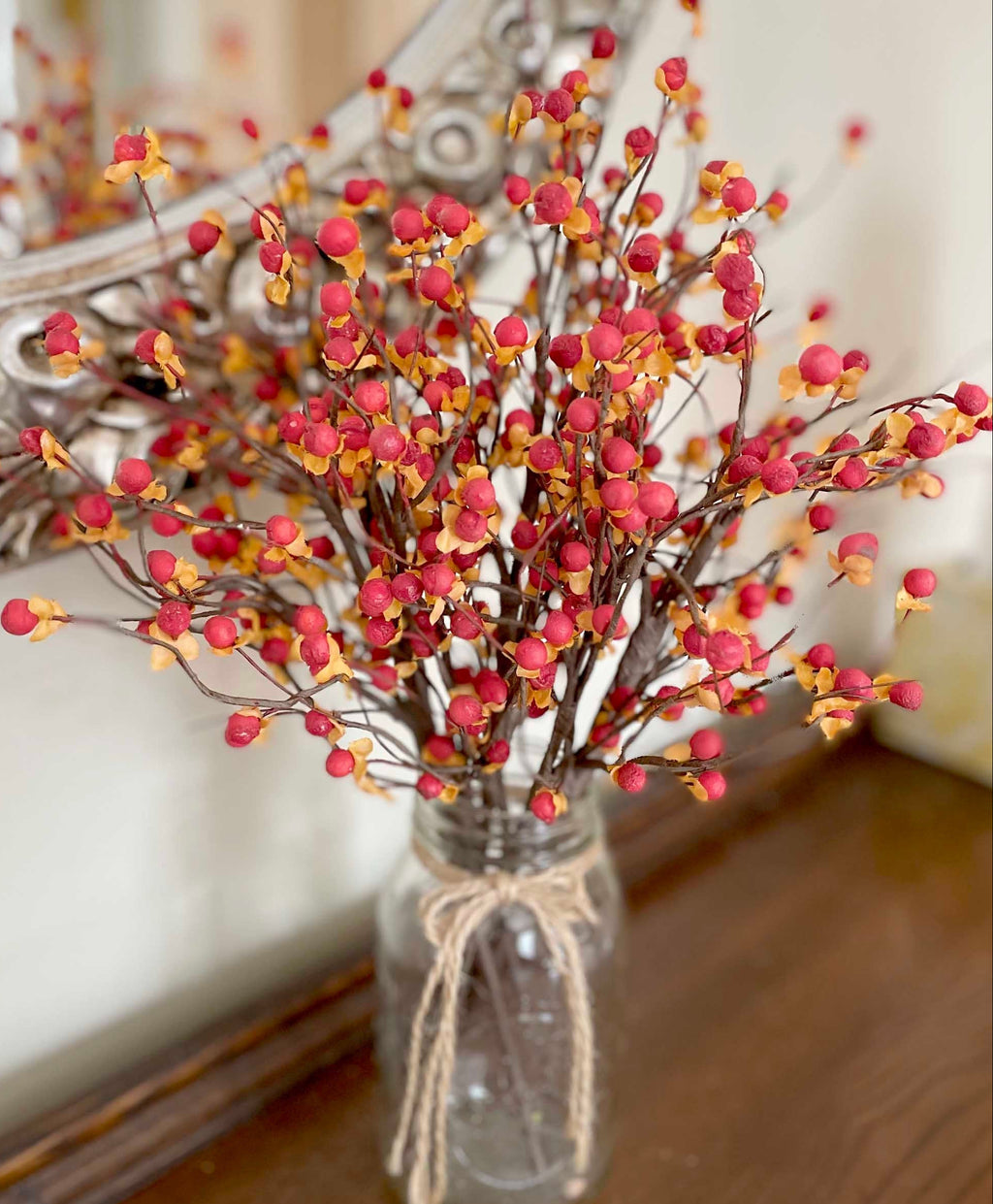 Decorative arrangement of red and orange berries in a glass vase with twine, against a neutral background.