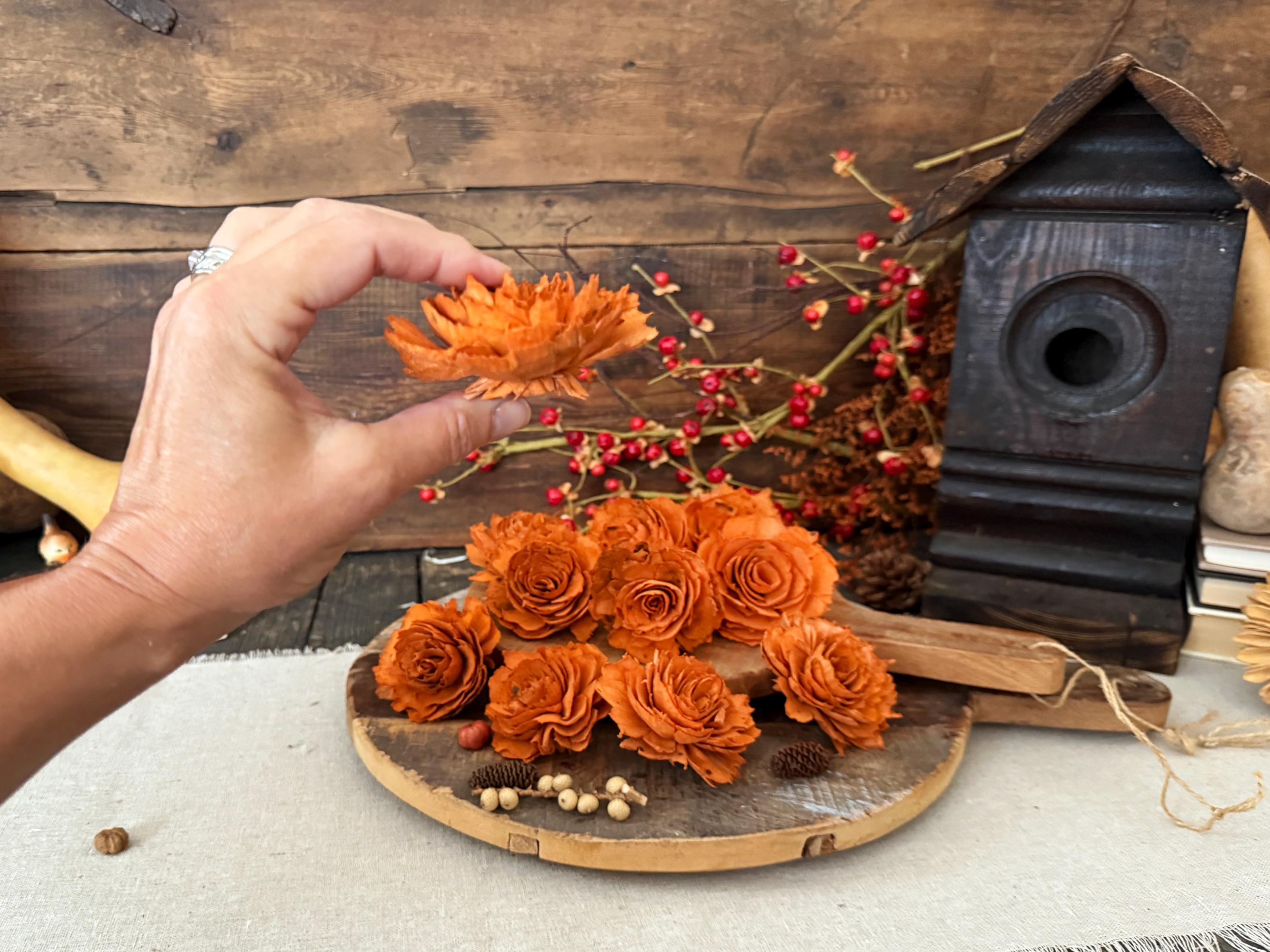 Hand holding orange artificial flowers to show profile. A wooden board with a rustic background hold s more flowers.