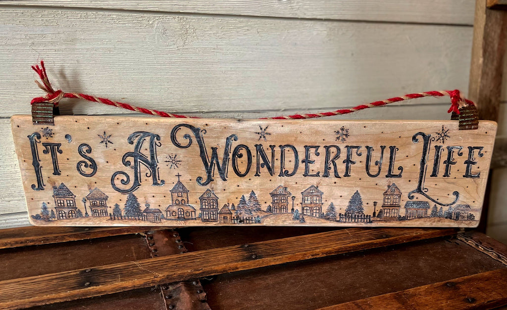 Wooden sign with 'It's a Wonderful Life' text and house illustrations on a rustic wooden surface.